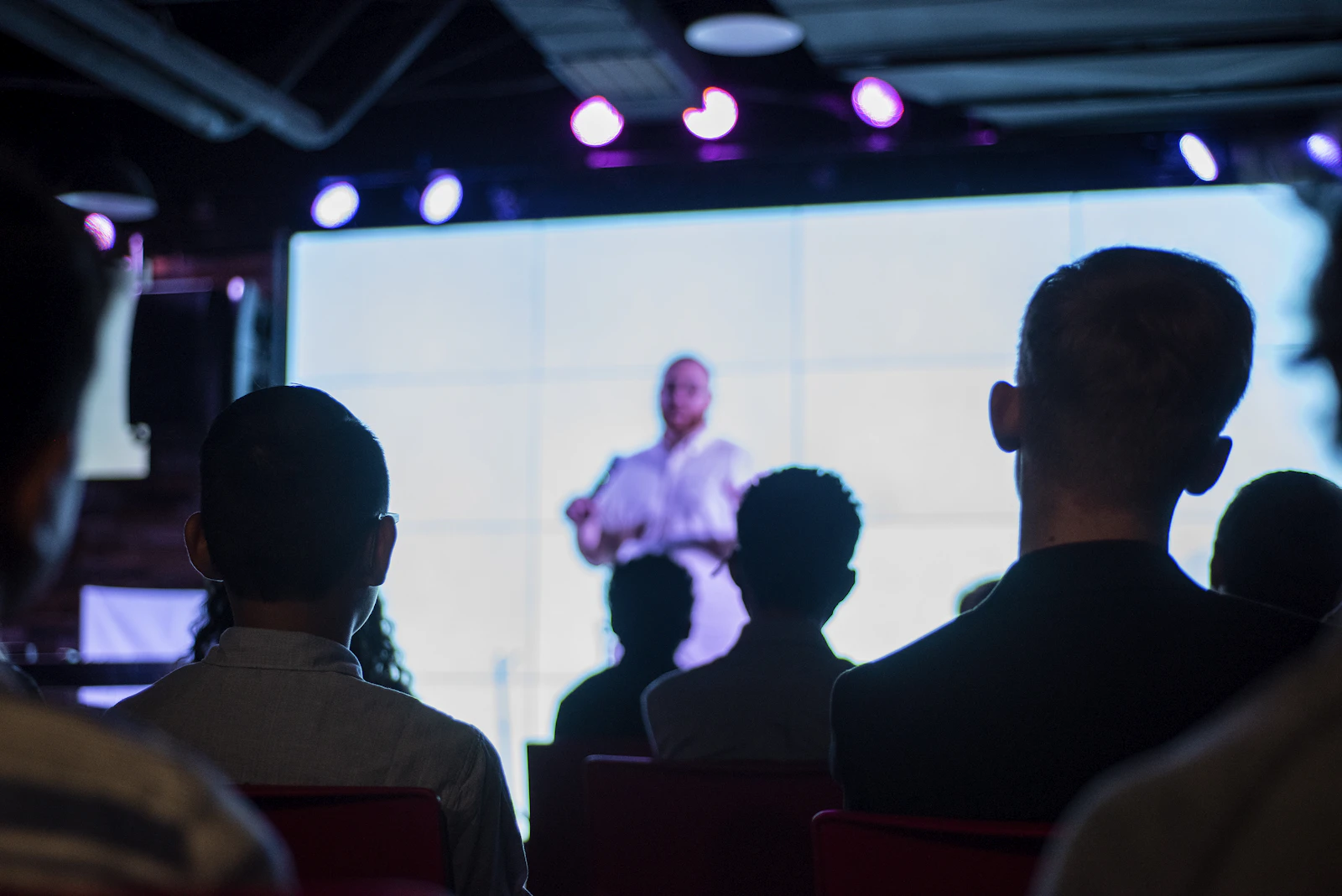 An audience watching a speaker on a bright screen in a dark room